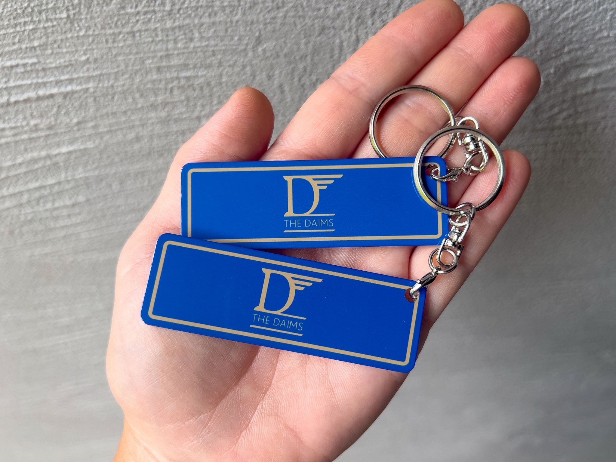 Two blue rectangular aluminium keychains with gold borders and a gold 'The Daims' logo, held in a person's hand against a grey textured wall.