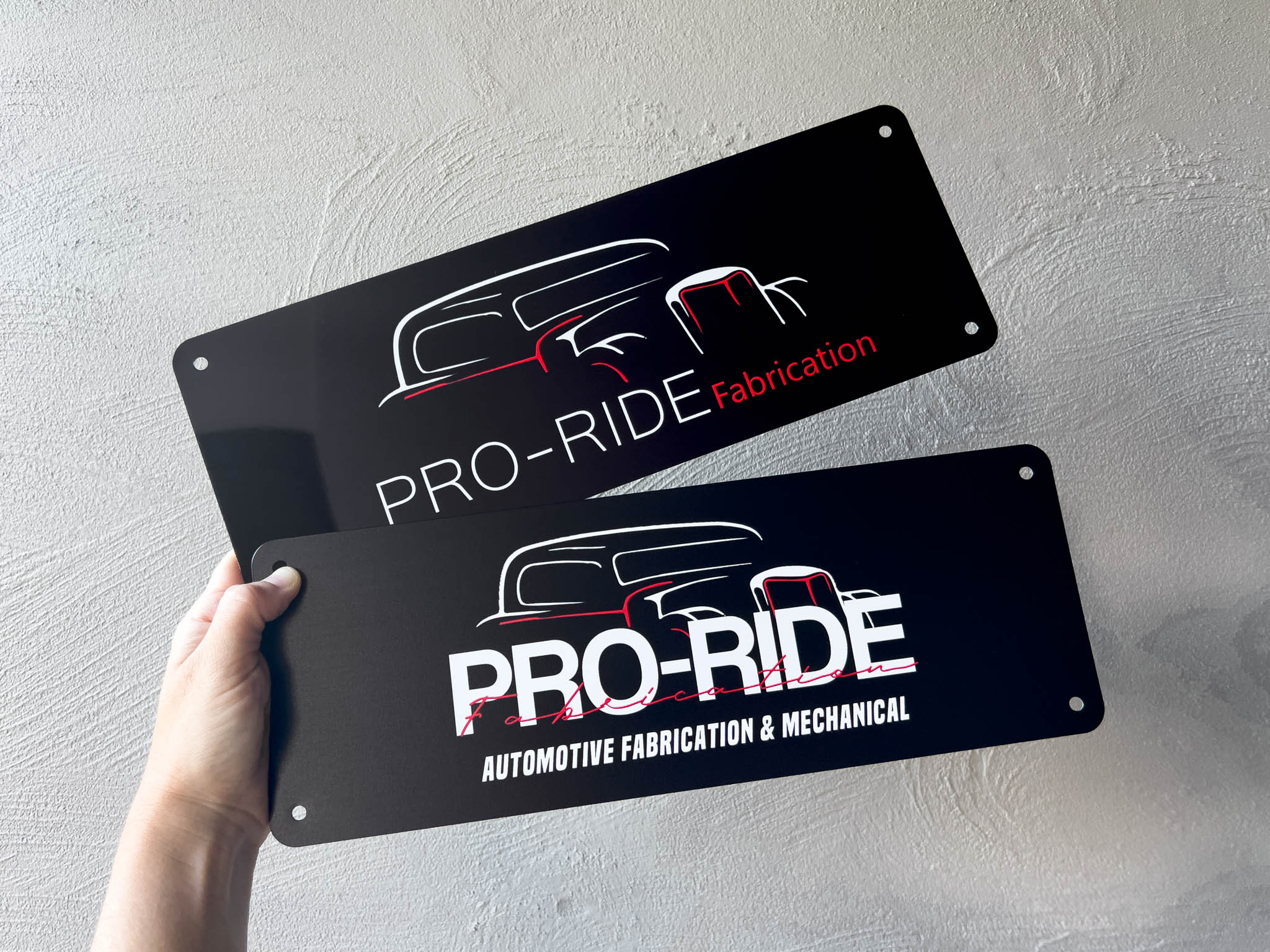 Two 'PRO-RIDE' branded metal logo number plates held against a light grey background.