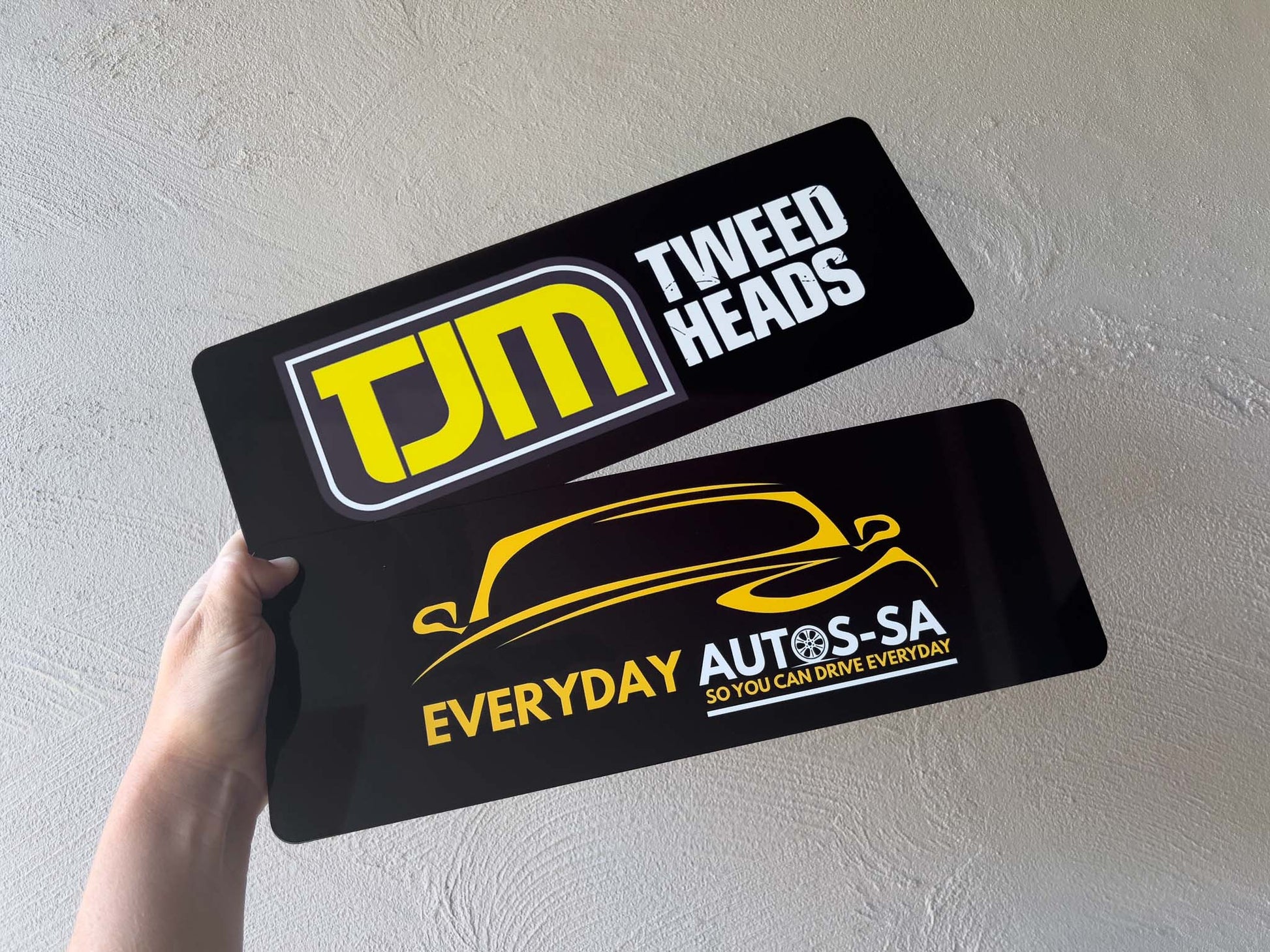 Two personalised logo plates with TJM and Everyday Autos-SA logos on a light grey background.