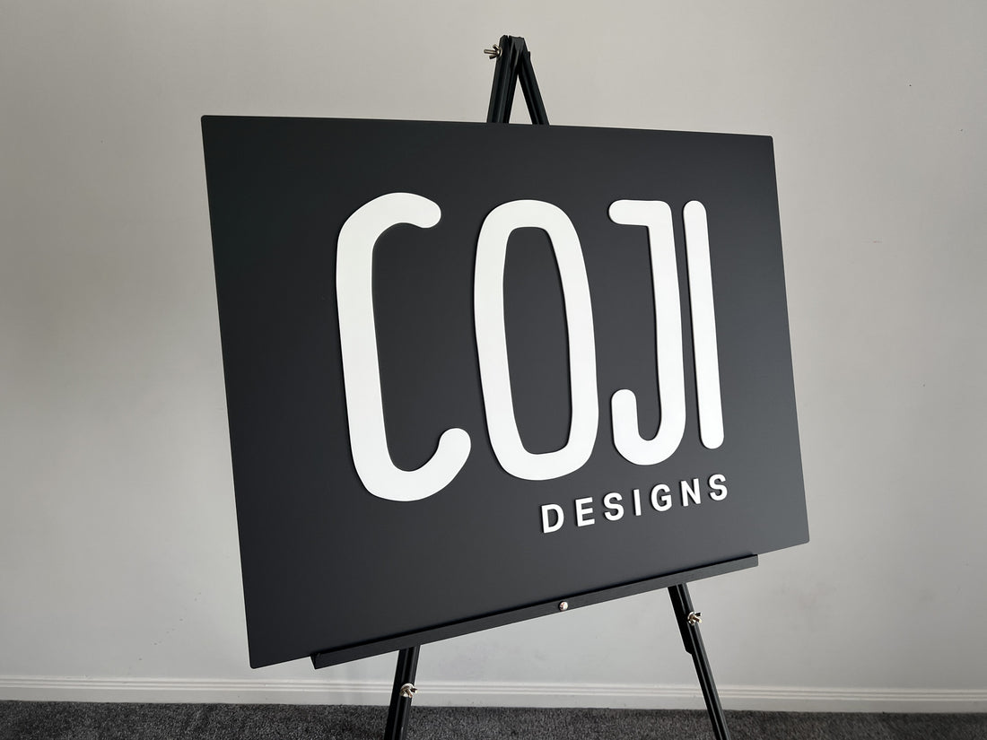 Black board with 'COJI DESIGNS' text on an easel against a gray wall.