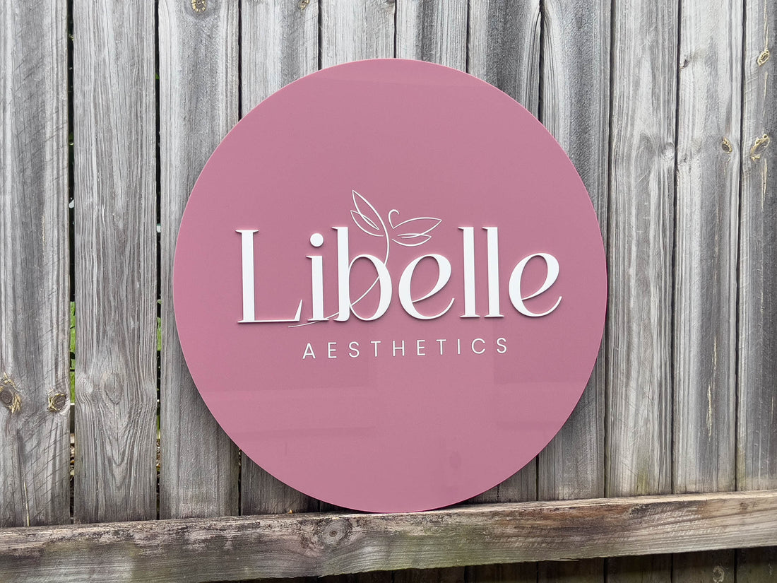 Round dusty pink acrylic sign with 'Libelle Aesthetics' logo on a wooden background