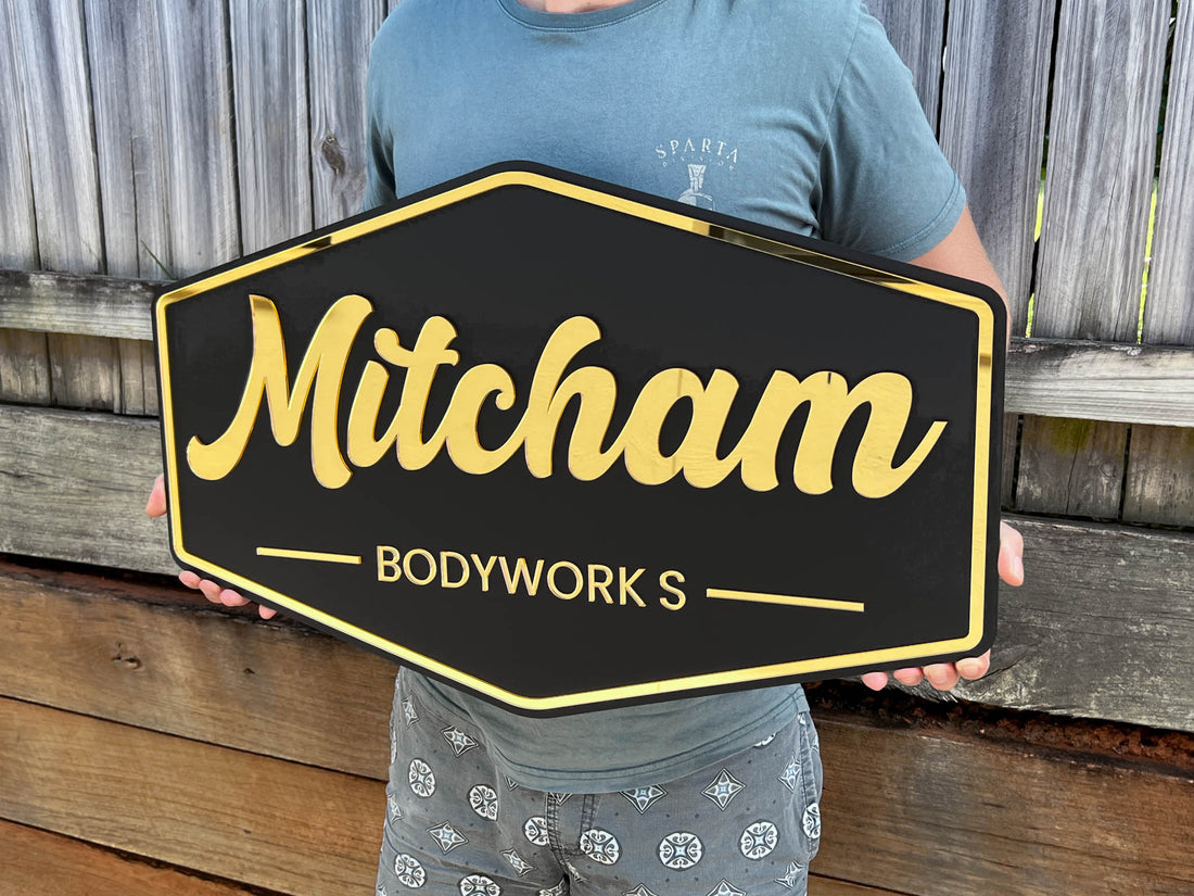 Person holding a black and gold sign with 'Mitcham Bodyworks' text against a wooden background