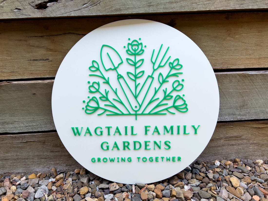Round white sign with green logo and text 'Wagtail Family Gardens Growing Together' on a wooden background.