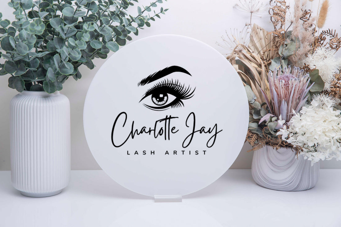 Round sign with 'Charlotte Jay Lash Artist' and an eye logo, placed on a white surface with decorative plants.