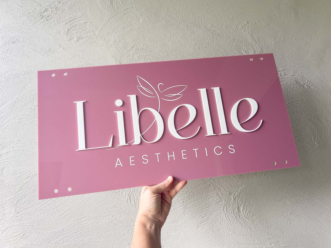 Hand holding a dusty pink acrylic logo sign with 'Libelle Aesthetics' on a light gray background