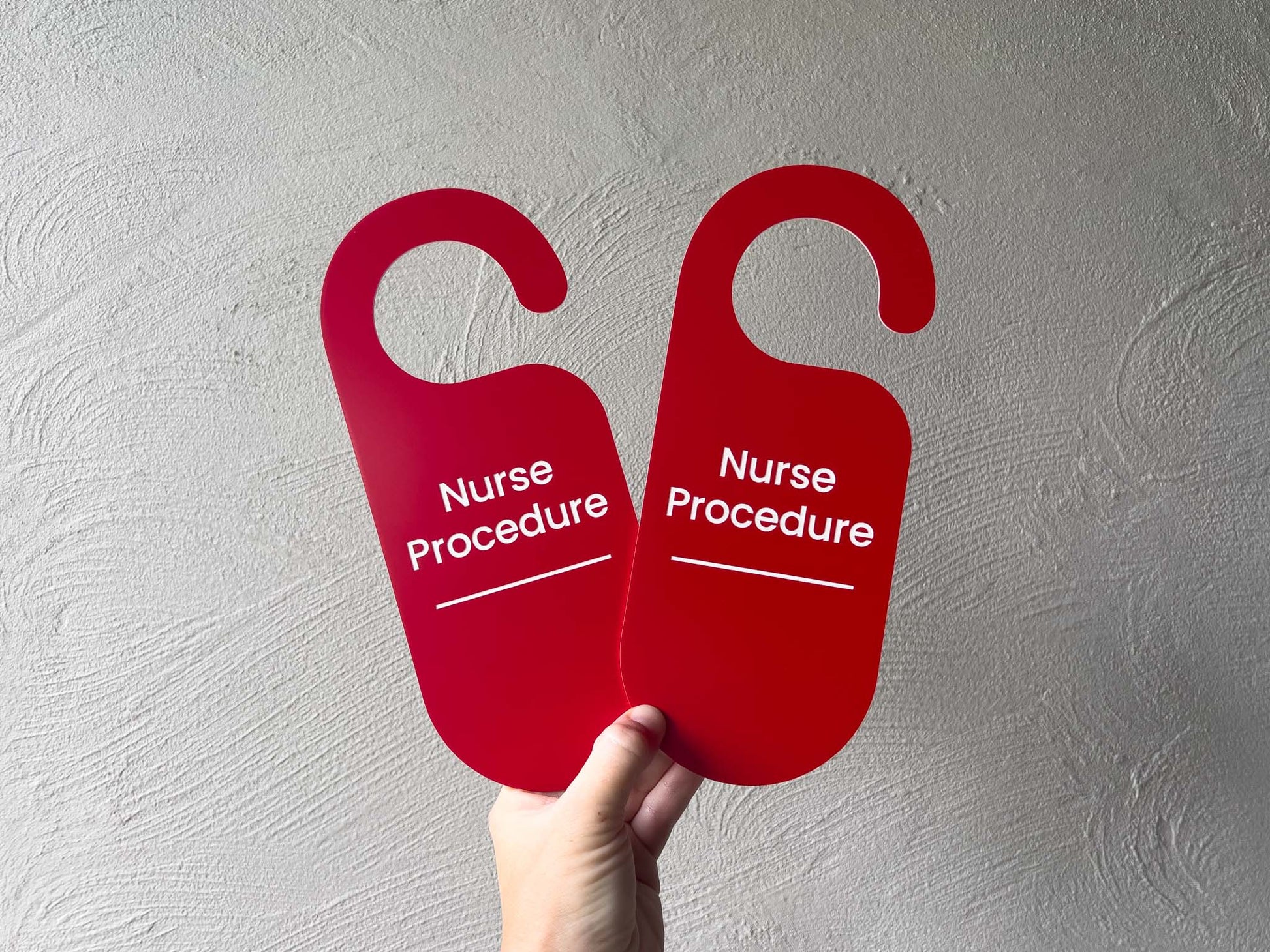 Two custom engraved red 'Nurse Procedure' door hangers with white text, held by a hand against a textured light grey wall.