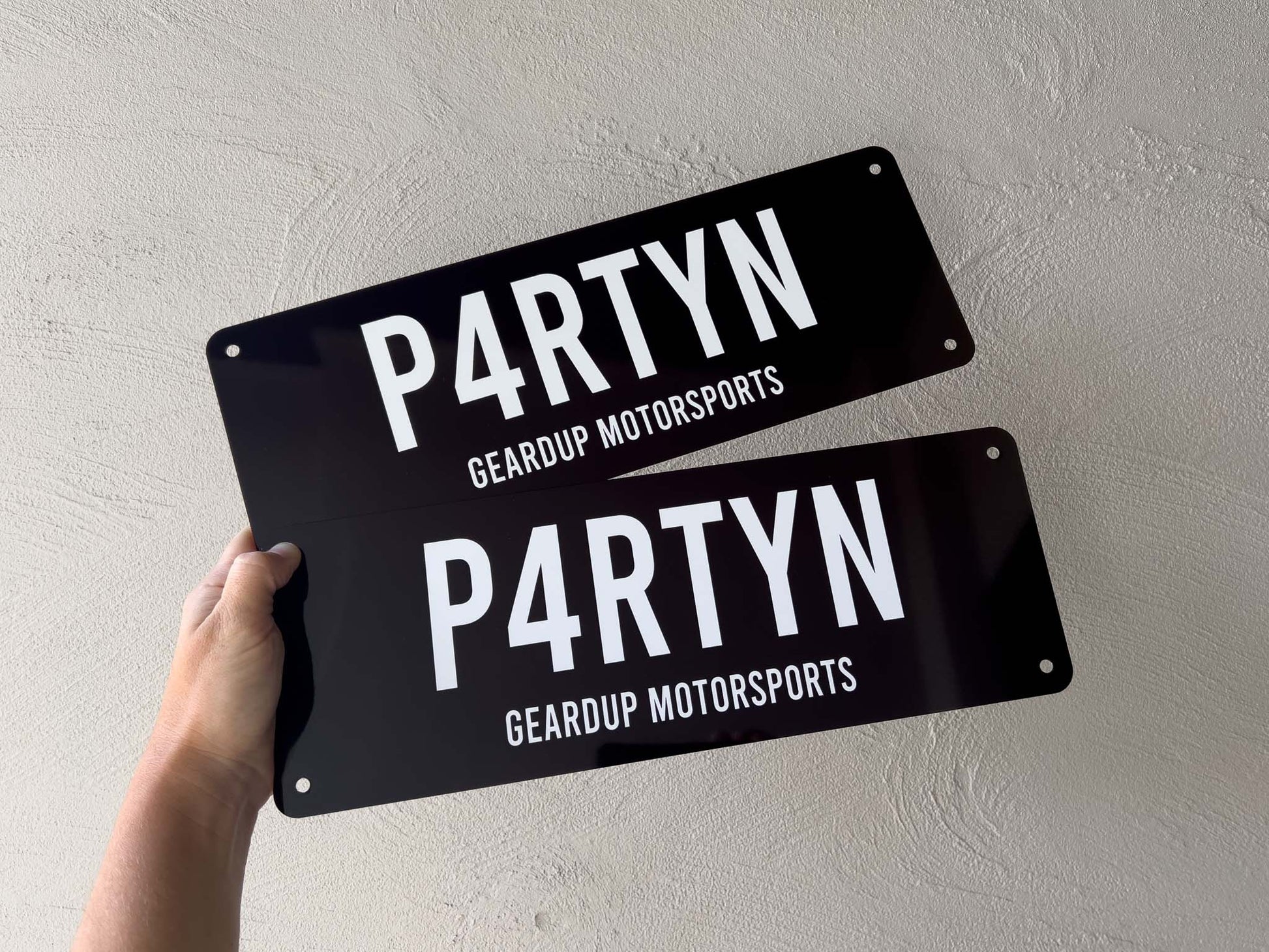 Two black number plate-style signs with 'PARTYN' and 'GEARDUP MOTORSPORTS' text held by a hand against a light grey background.