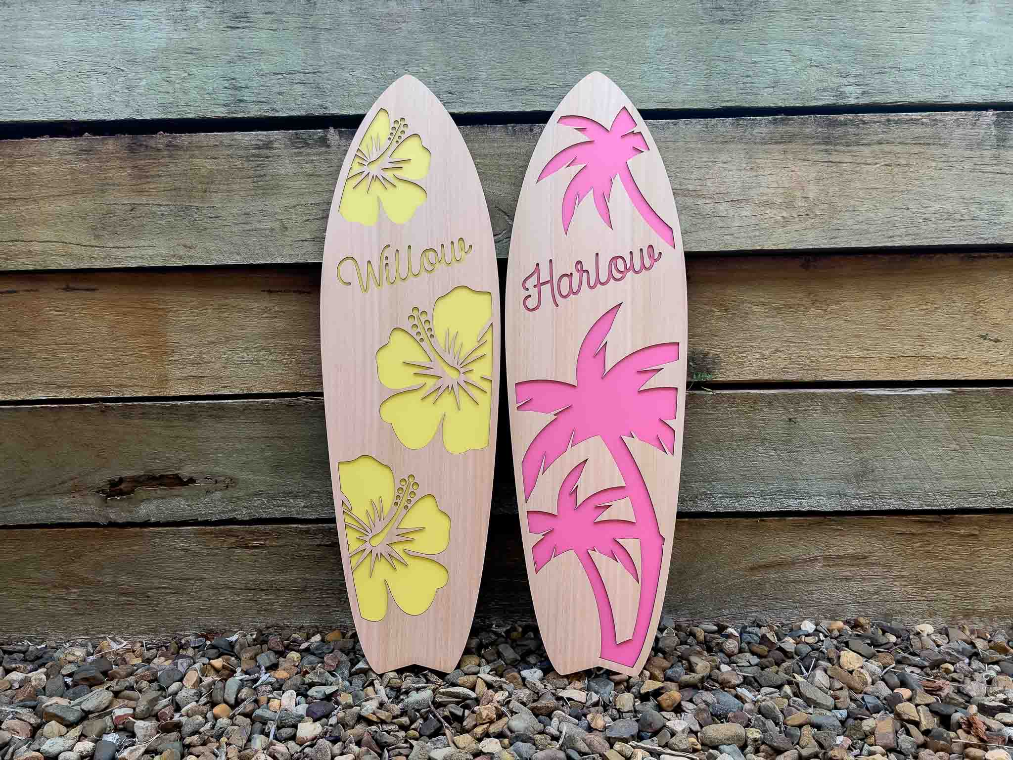 Two wooden surfboards with 'Willow' in lemon yellow and 'Harlow' in pink.