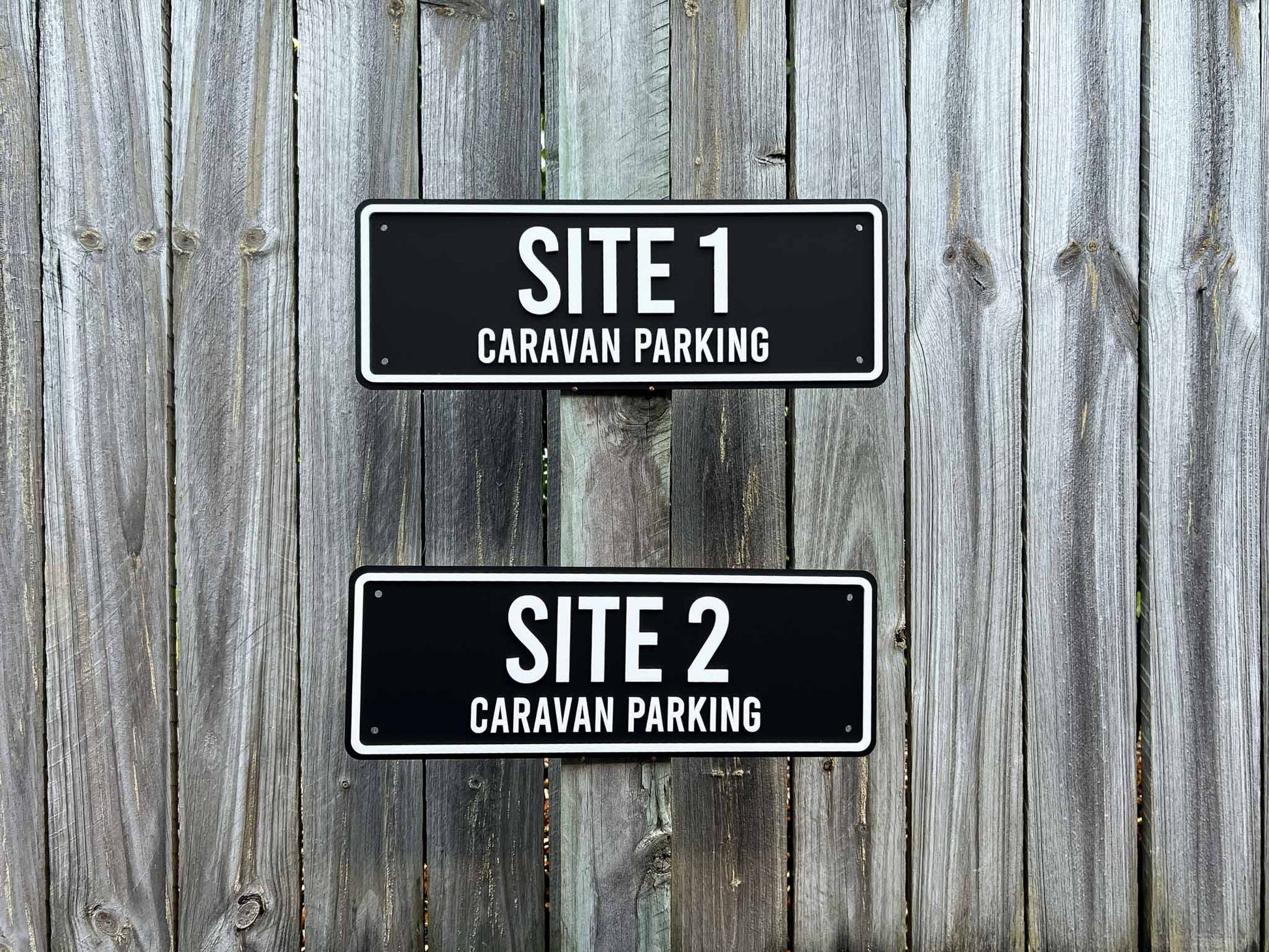 Two outdoor black and white signs indicating 'SITE 1 CARAVAN PARKING' and 'SITE 2 CARAVAN PARKING' on a wooden fence.
