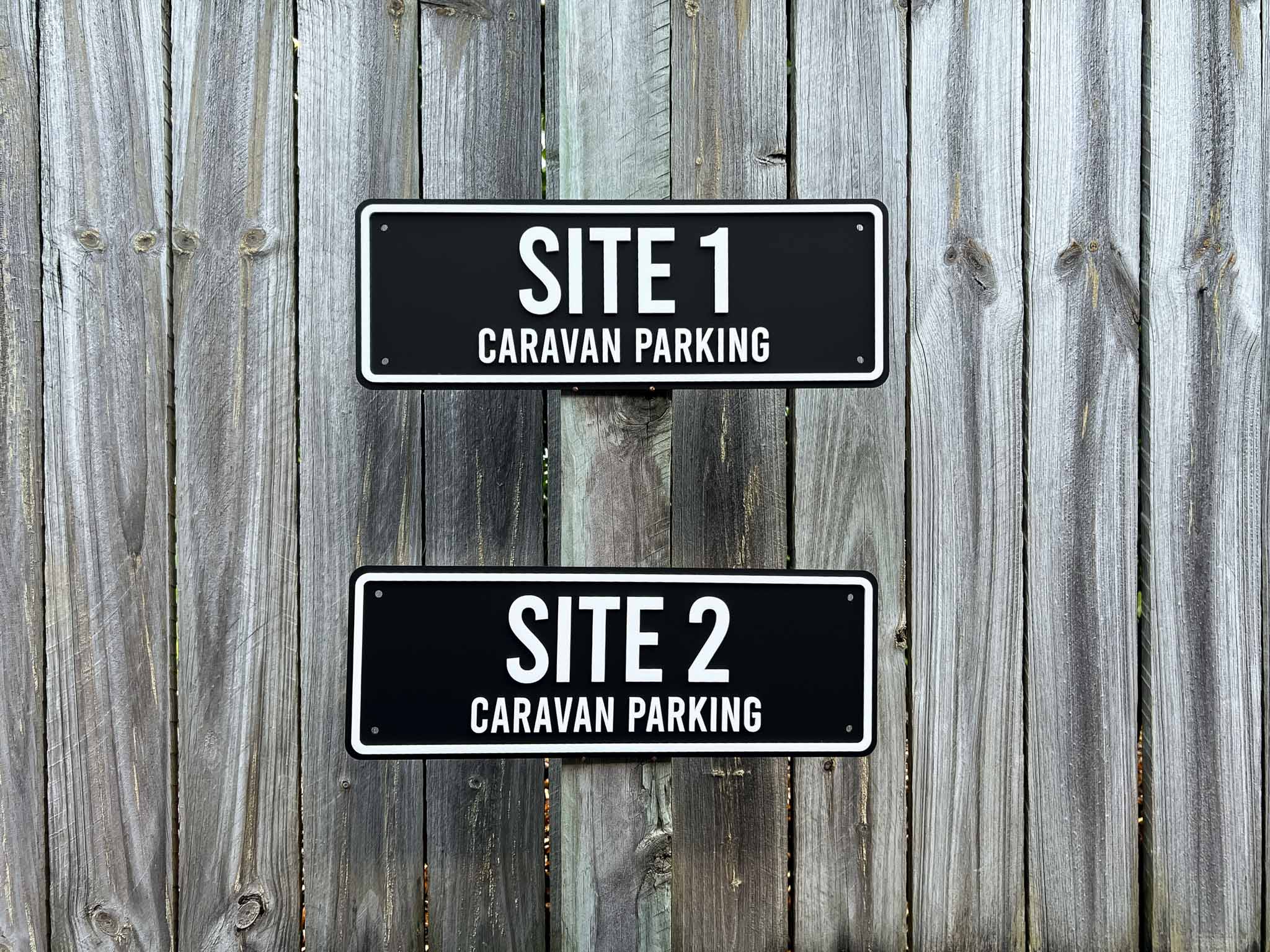 Two outdoor black and white signs indicating 'SITE 1 CARAVAN PARKING' and 'SITE 2 CARAVAN PARKING' on a wooden fence.