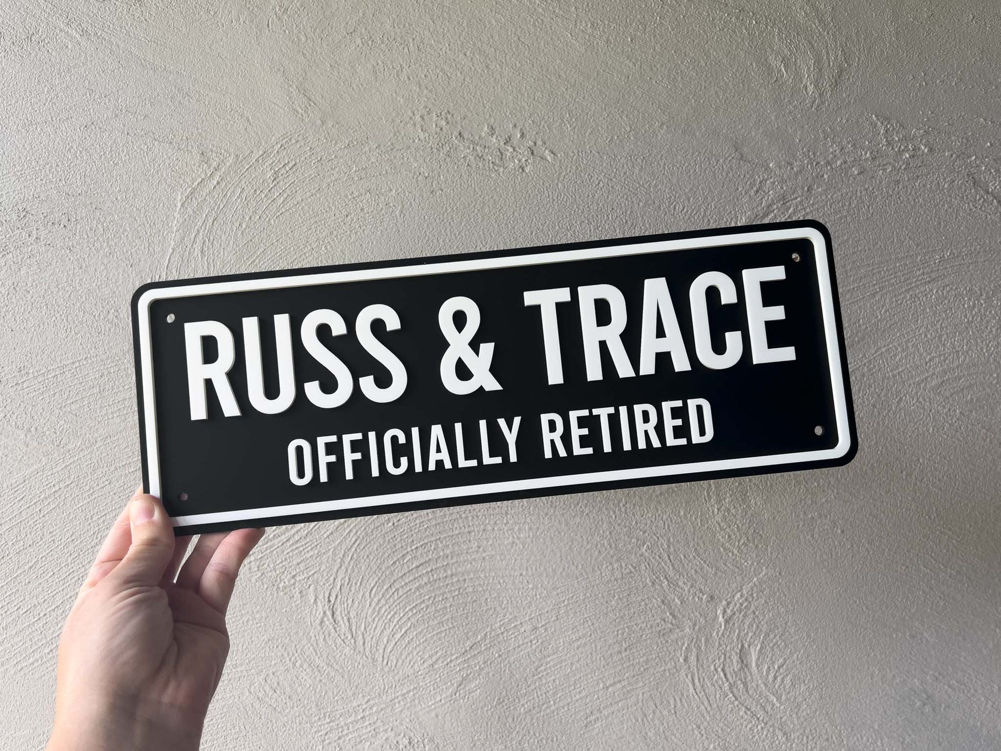 Hand holding a black and white acrylic plate sign with 'Russ & Trace Officially Retired' text against a plain background