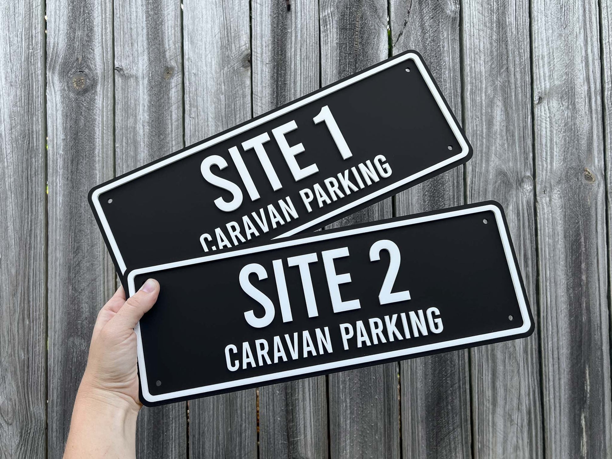 Two black signs with white text indicating 'Site 1 Caravan Parking' and 'Site 2 Caravan Parking' held against a wooden background.