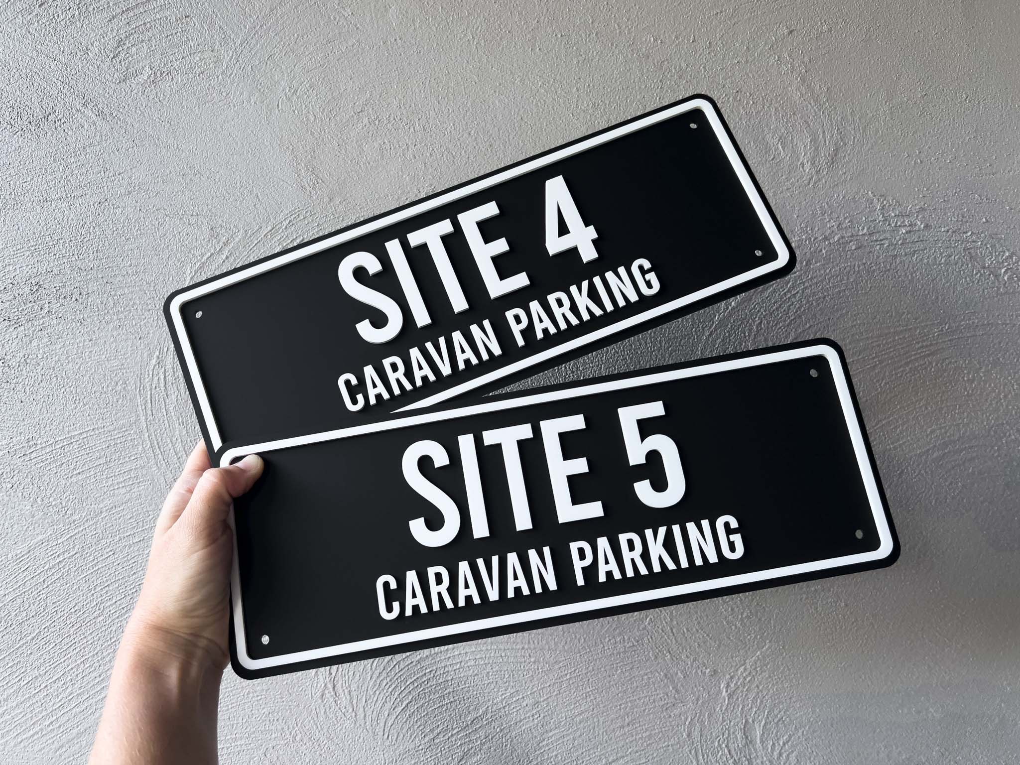 Two outdoor black signs with white text indicating 'SITE 4 CARAVAN PARKING' and 'SITE 5 CARAVAN PARKING' held against a grey background.