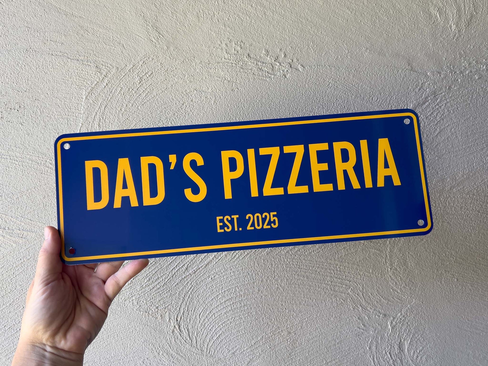 Hand holding a blue and yellow sign with 'DAD'S PIZZERIA EST. 2025' text against a light grey wall.