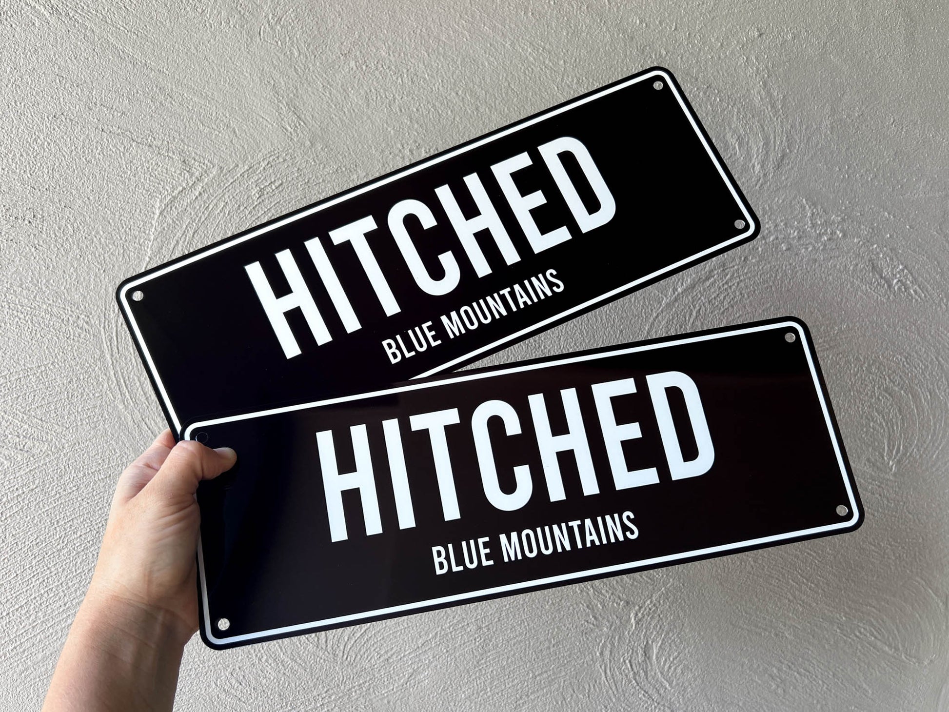Two black signs with 'Hitched Blue Mountains' text held against a light grey background