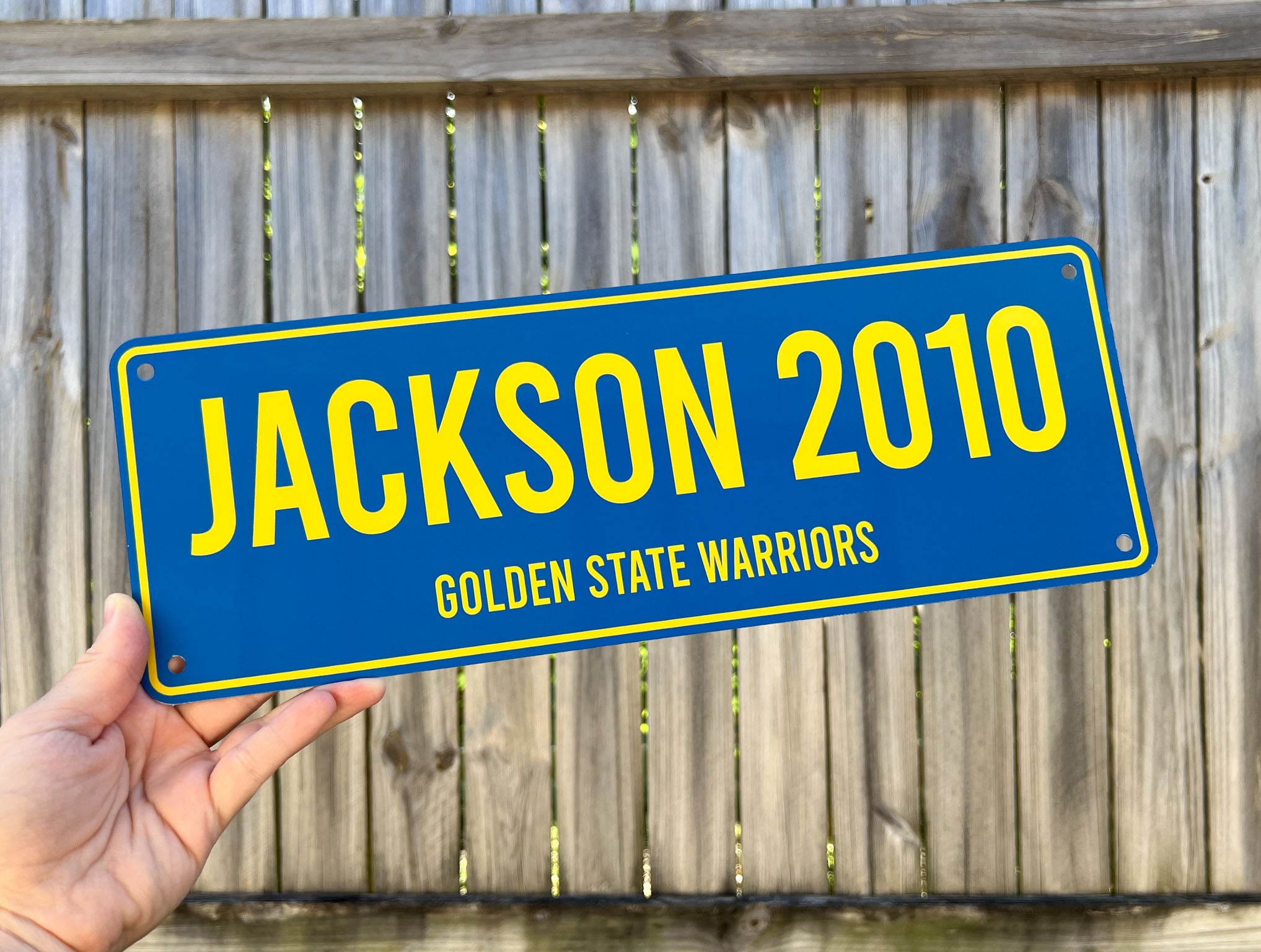 Hand holding a blue and yellow sign with 'Jackson 2010 Golden State Warriors' text against a wooden fence background.