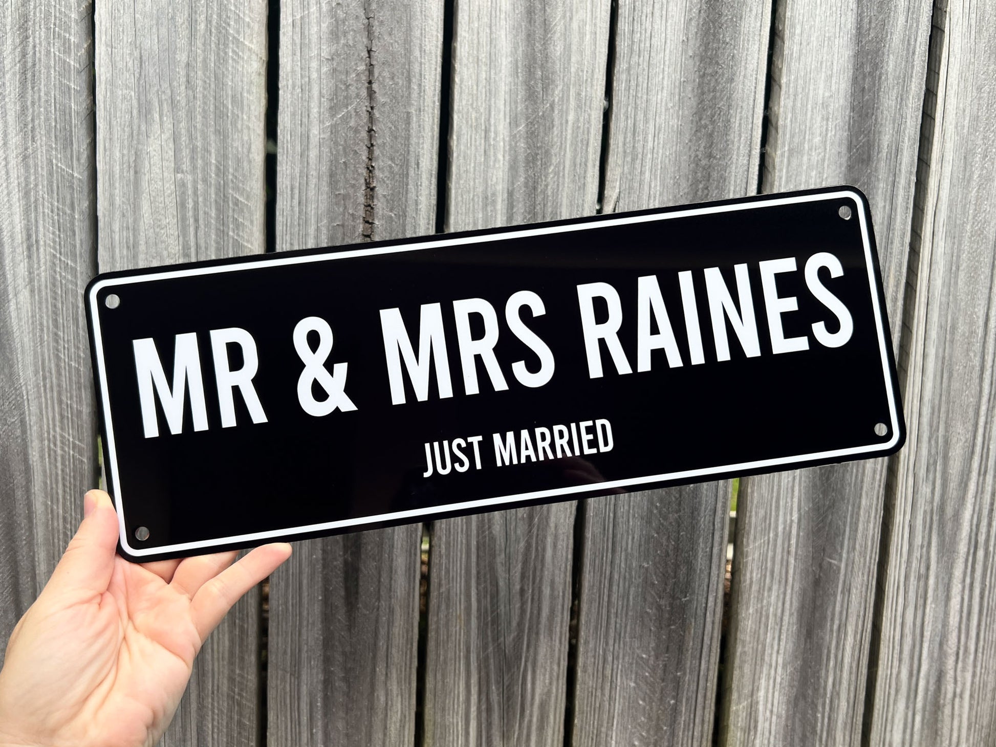 Hand holding a black sign with 'MR & MRS RAINES JUST MARRIED' text against a wooden background