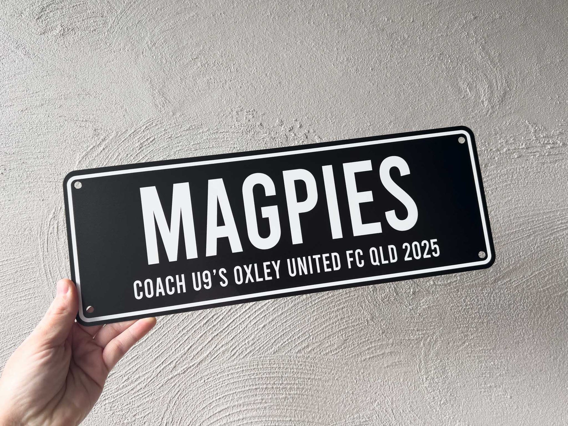 Hand holding a black sign with 'MAGPIES' and sports team details against a light grey background