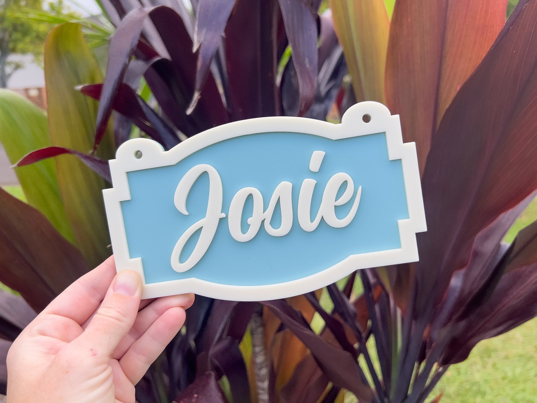 Hand holding a custom sign with 'Josie' in a dusty coastal blue and beige colour against a garden background