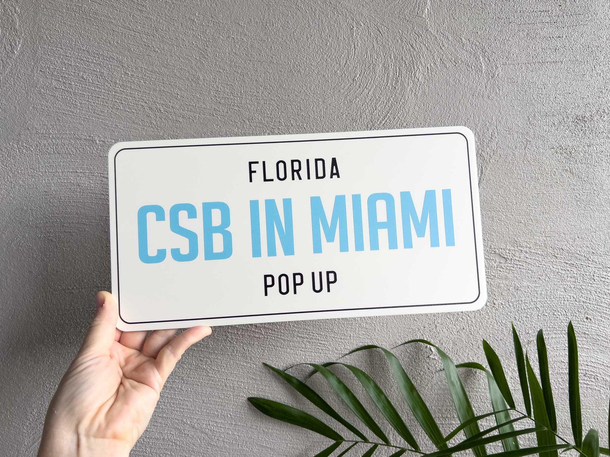 Hand holding a sign with 'Florida CSB IN MIAMI POP UP' text against a grey background.