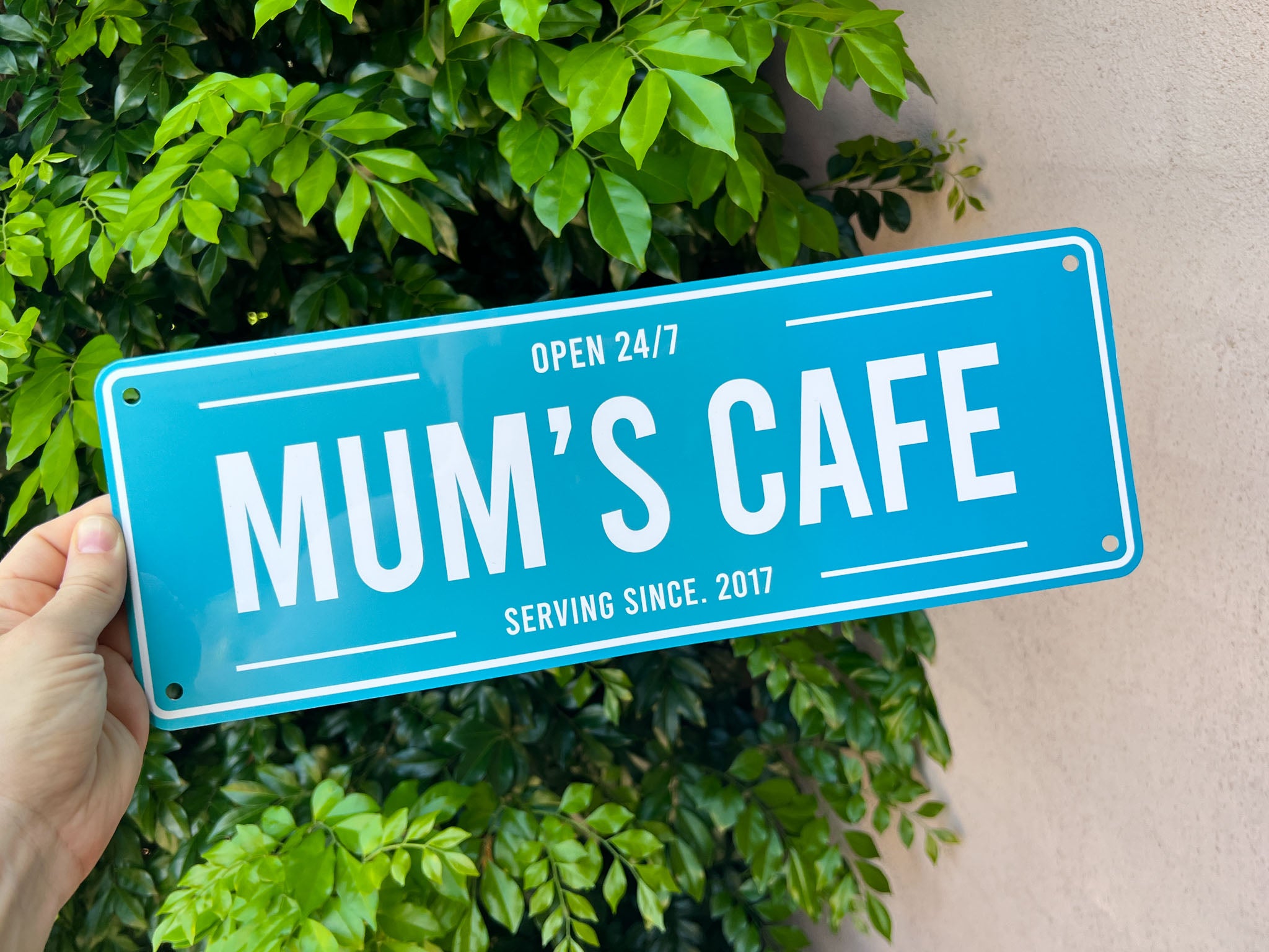 Blue custom printed sign with 'Mum's Cafe' text held by a hand against a green leafy background