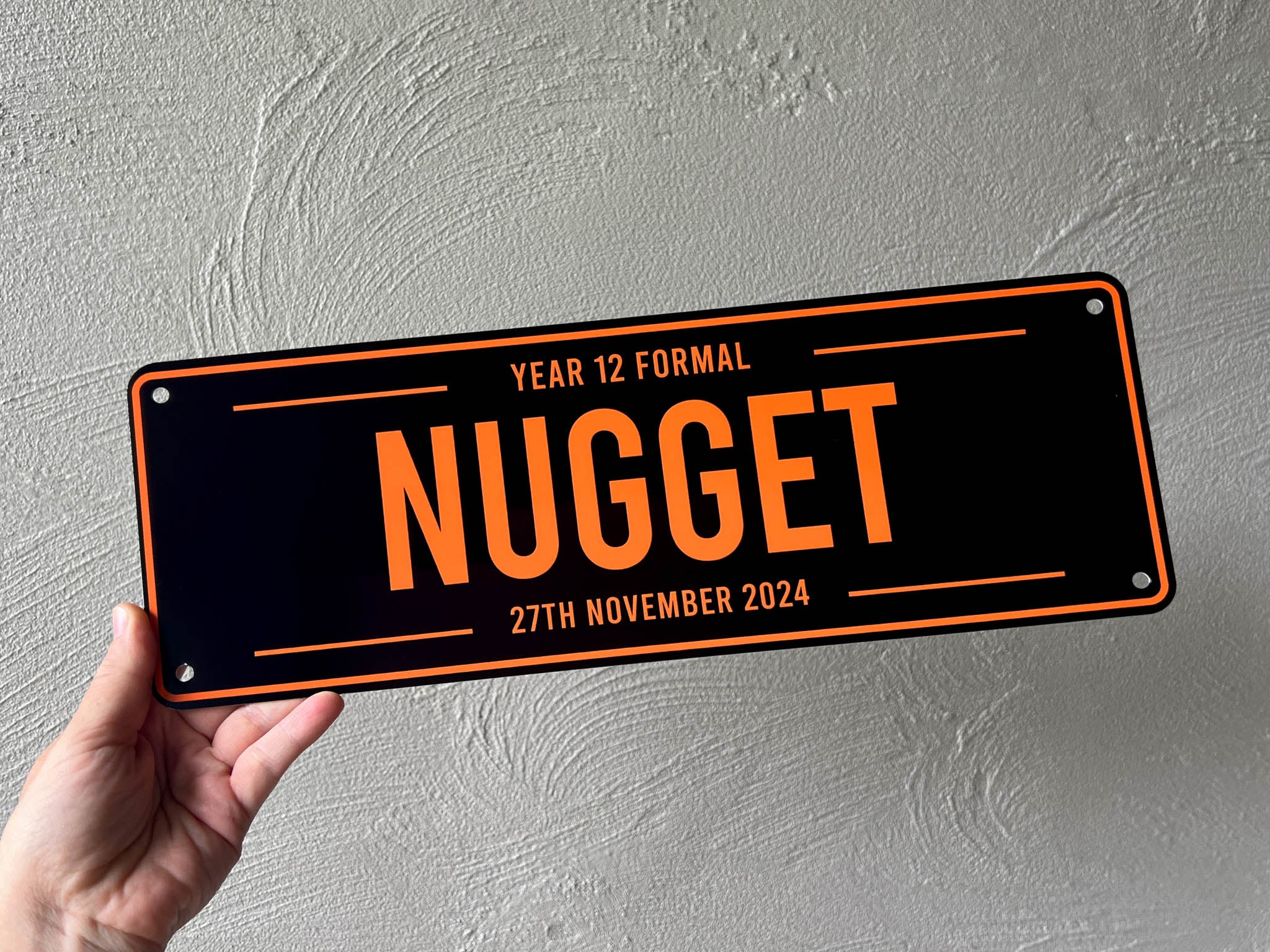 Hand holding a metal plate with 'NUGGET' and year 12 formal event details against a grey background