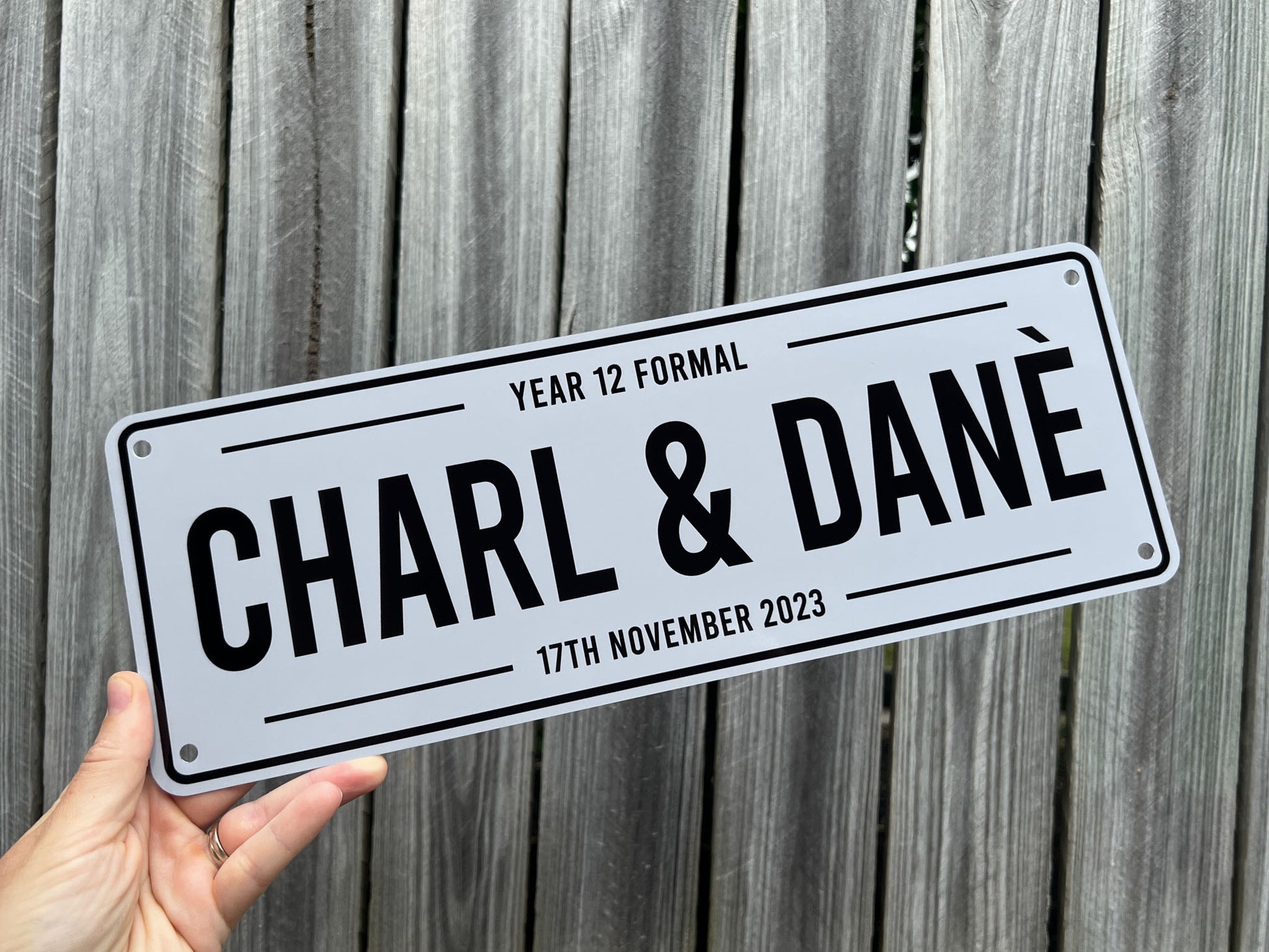 Custom metal car plate with 'CHARL & DANE Year 12 Formal' text held against a wooden background