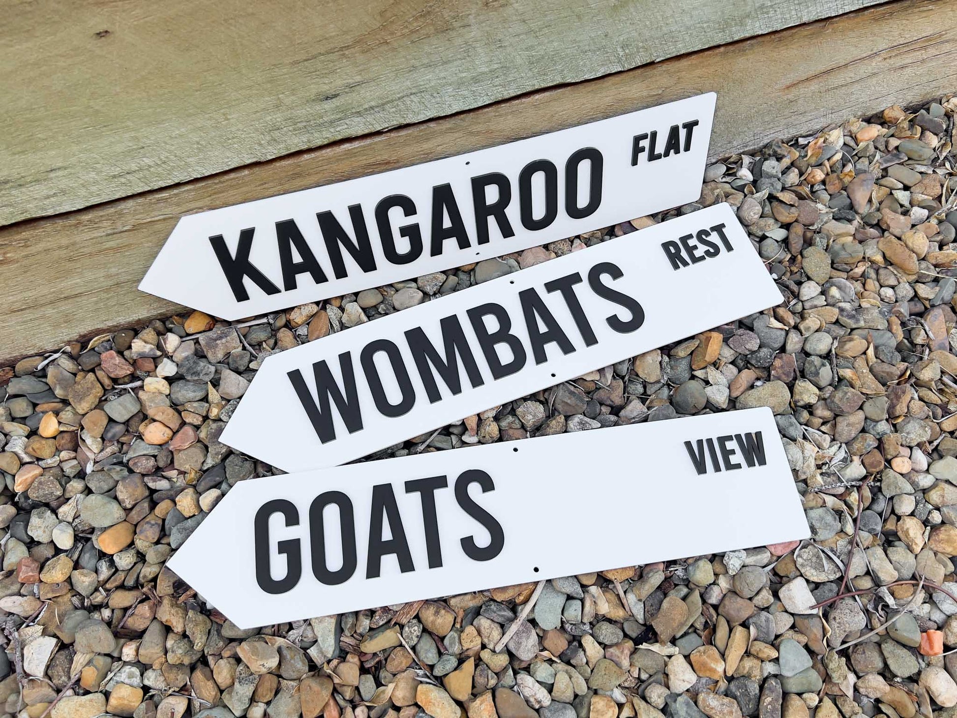 Three white signs that say 'KANGAROO FLAT', 'WOMBAT'S REST' & 'GOATS VIEW' with black text on a gravel surface
