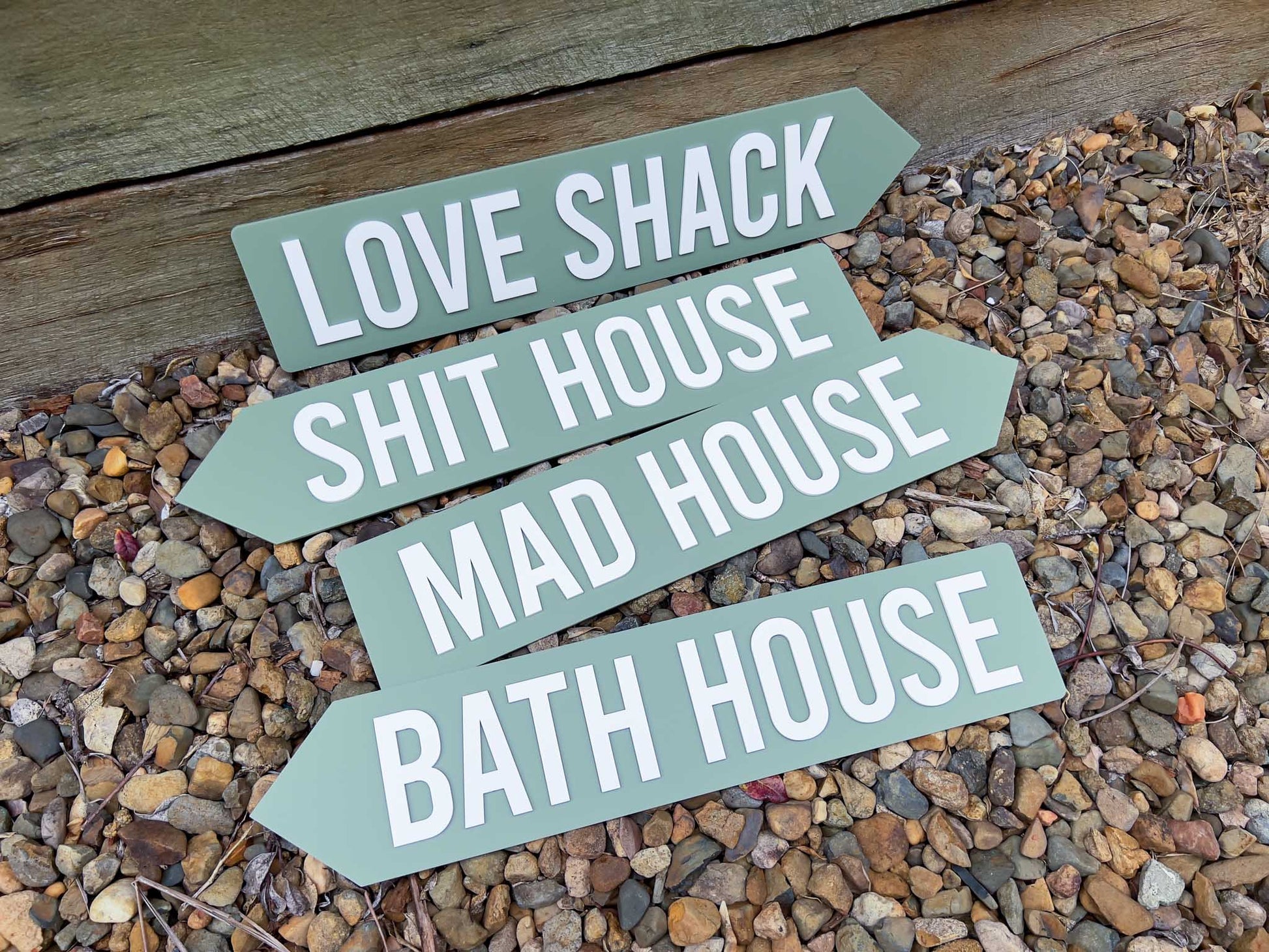 Four green signs that read 'LOVE SHACK', 'SHIT HOUSE', 'MAD HOUSE' & 'BATH HOUSE' with white text on a gravel surface