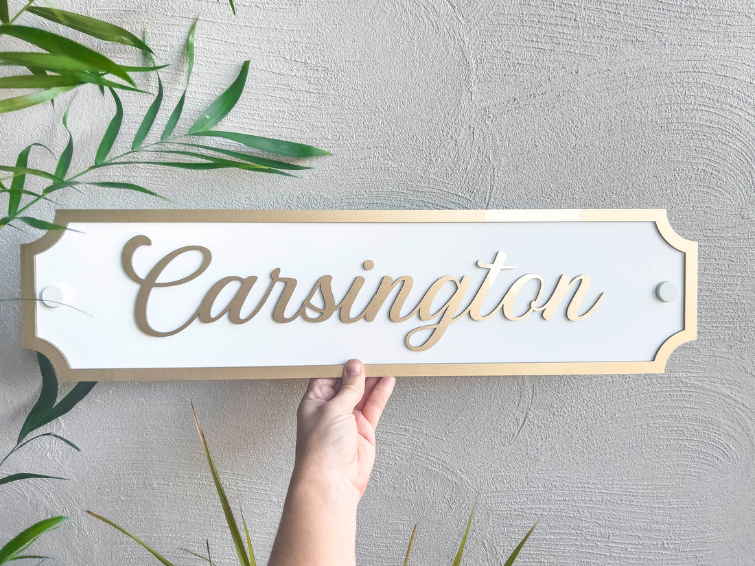 A stunning metallic gold and white tropical 3D acrylic panel that reads 'Carsington' in metallic gold and white held next to a palm tree.