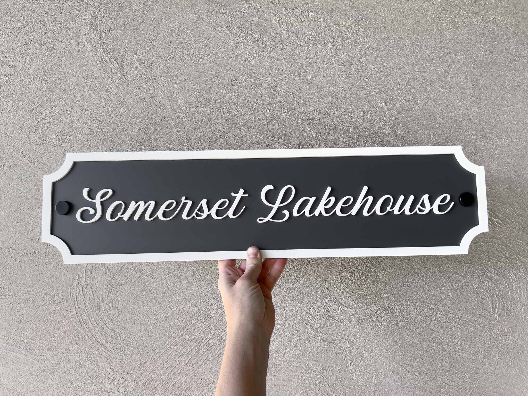 Hand holding a custom made sign with 'Somerset Lakehouse' text in dark grey and white against a plain background